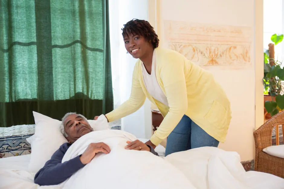Caregiver kneeling beside an elderly resident in an armchair in a softly lit aged care bedroom with family photos and a bedside call point