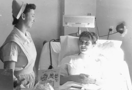 Nurse and care assistant responding at a patient's bedside in a modern hospital room, with a wall-mounted nurse call station beside the bed